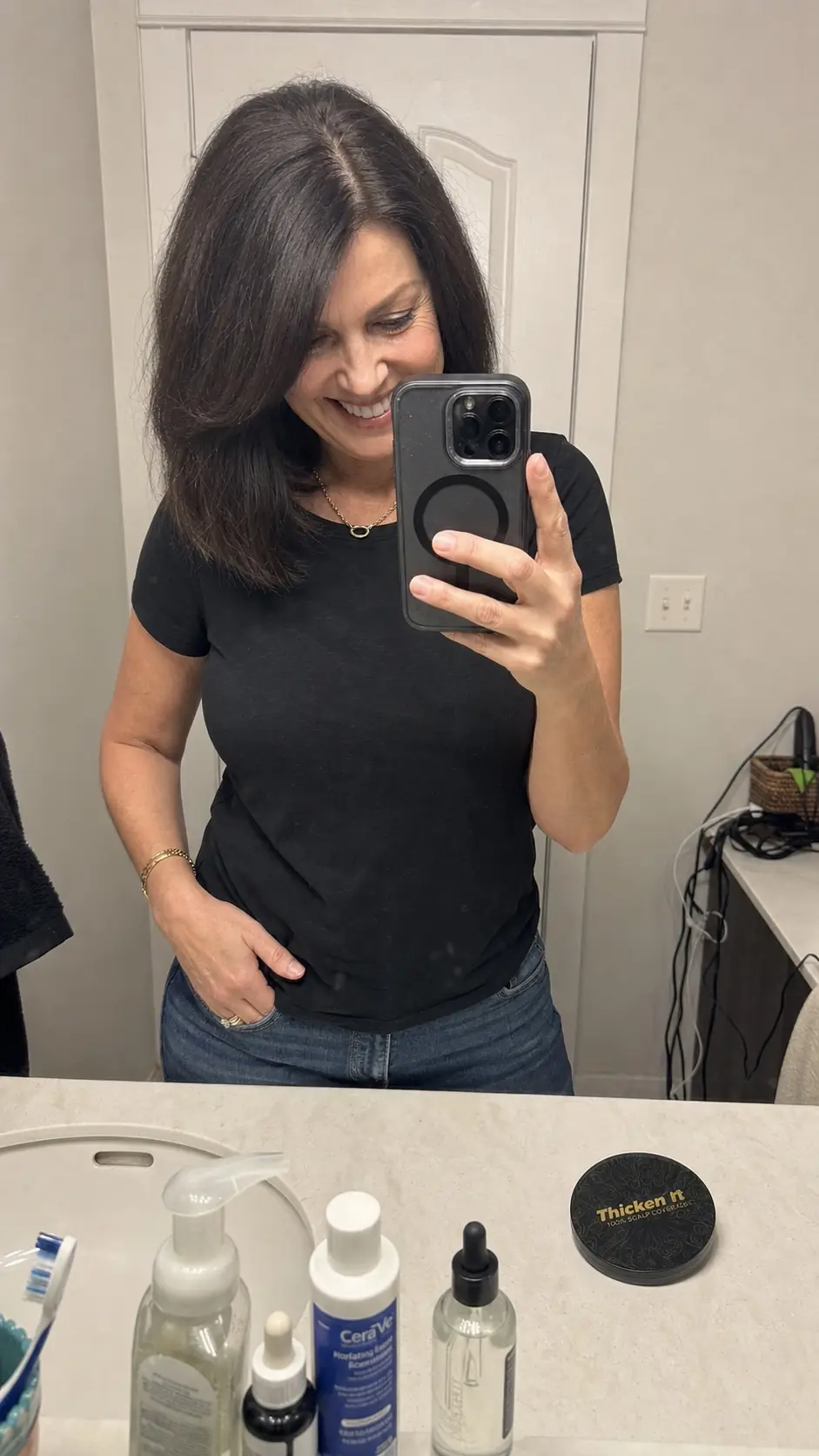 Smiling woman with dark shoulder-length hair holding her phone for a mirror selfie, part line looking full and natural